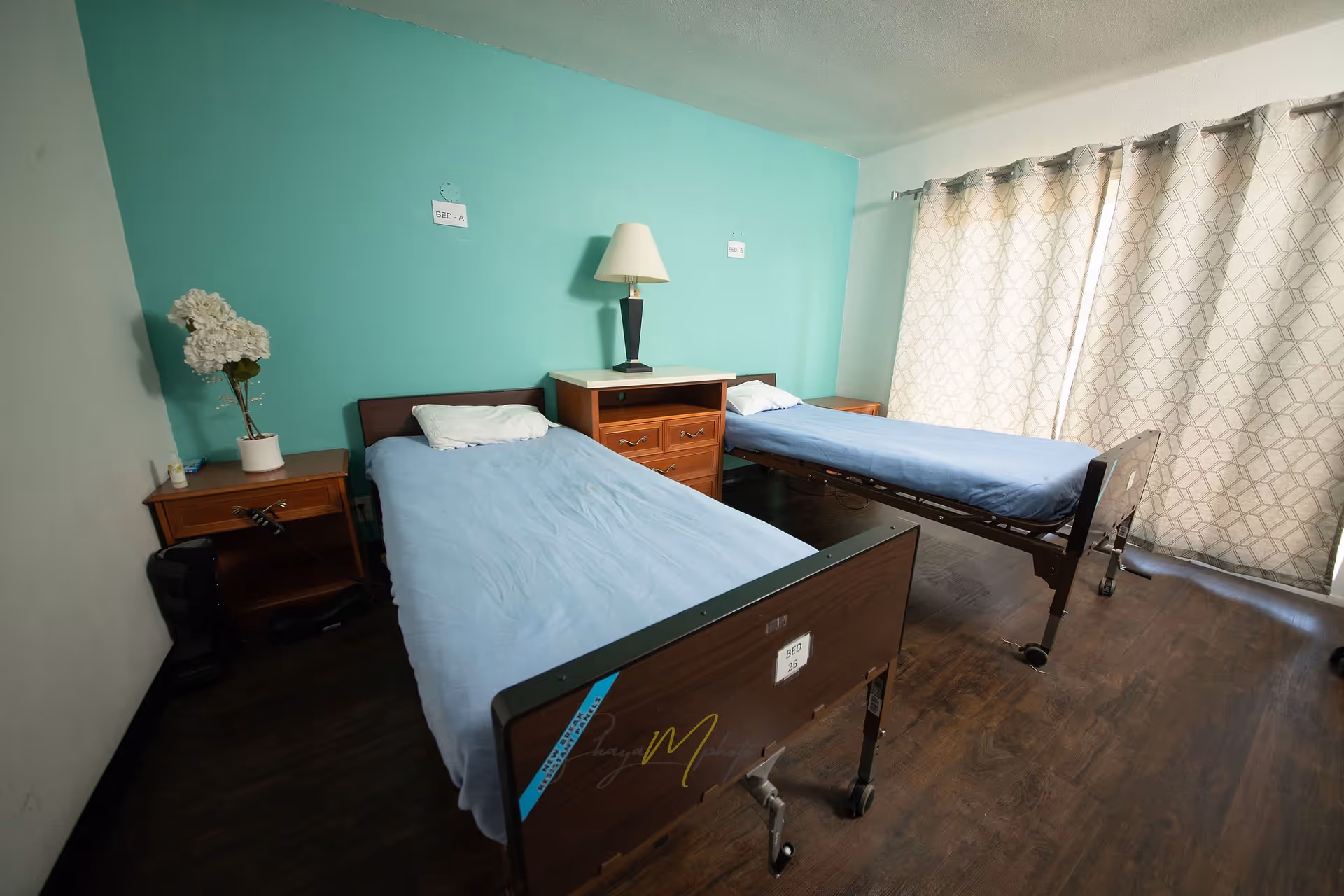 A bedroom in an assisted living facility with two single beds, each with a blue sheet and white pillow. Between the beds is a wooden nightstand with a lamp on top. The wall behind the beds is painted teal, and there are labels above each bed reading 'BED - A' and 'BED - B'. A window with patterned curtains is on the right side of the room, and there is a small table with a white flower arrangement on the left side.