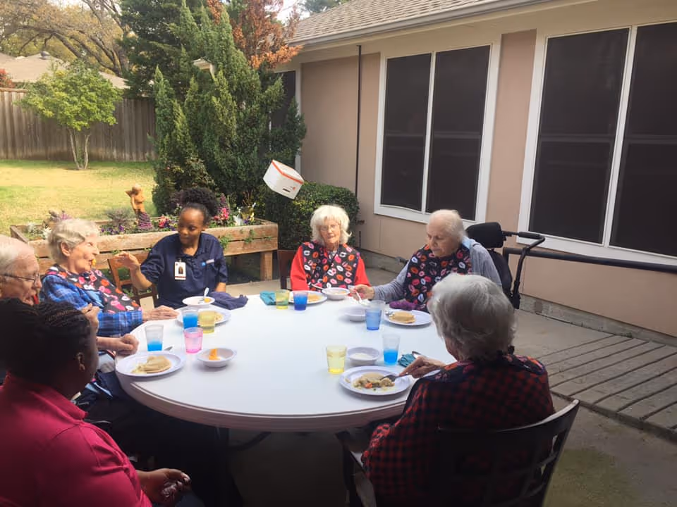 A caregiver and several elderly residents sit around a round outdoor table on a patio eating a meal.