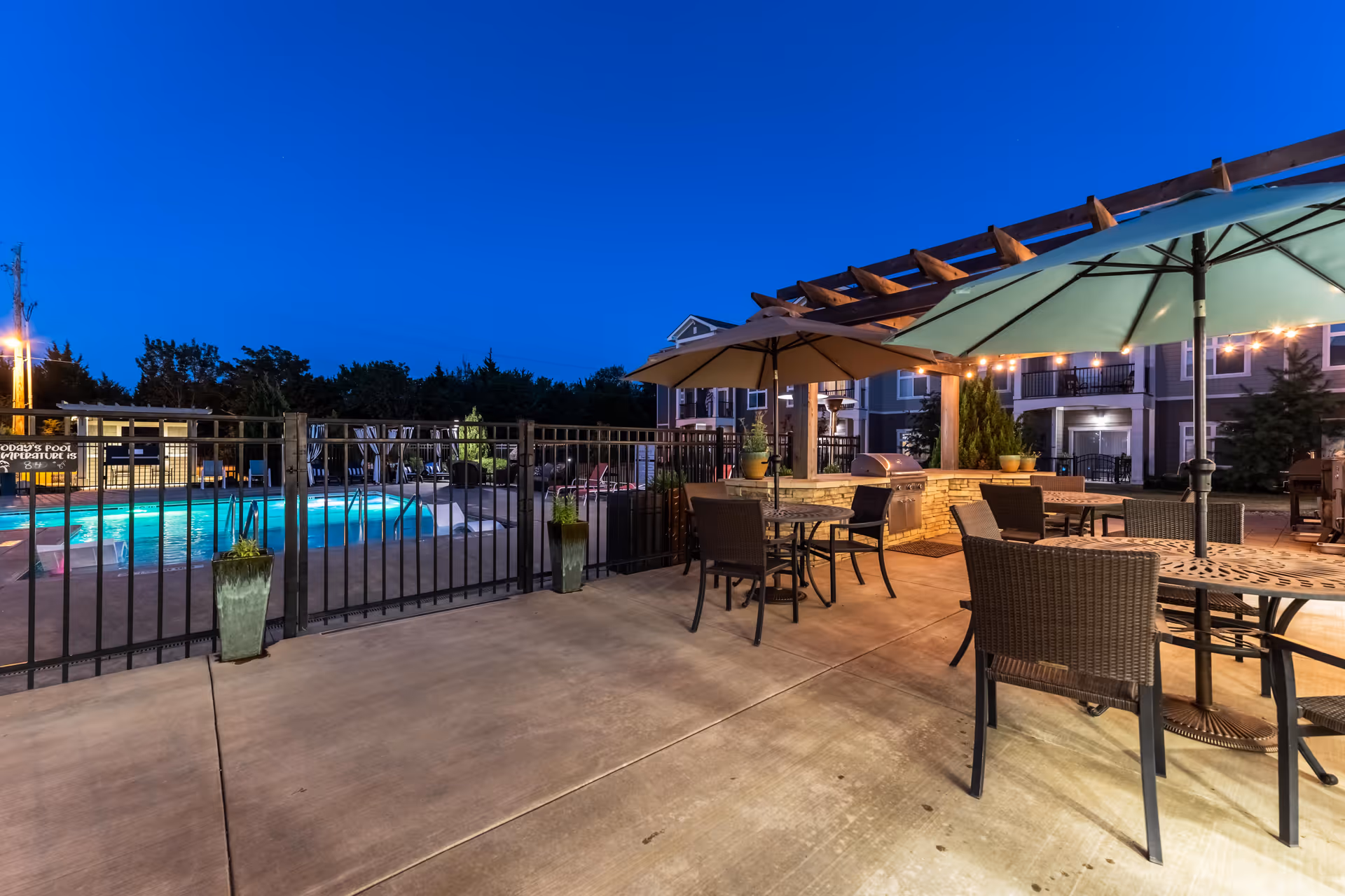 Outdoor patio area at dusk with tables, chairs, and large umbrellas under a wooden pergola. A fenced swimming pool with glowing blue water is visible in the background, along with residential buildings and some greenery.