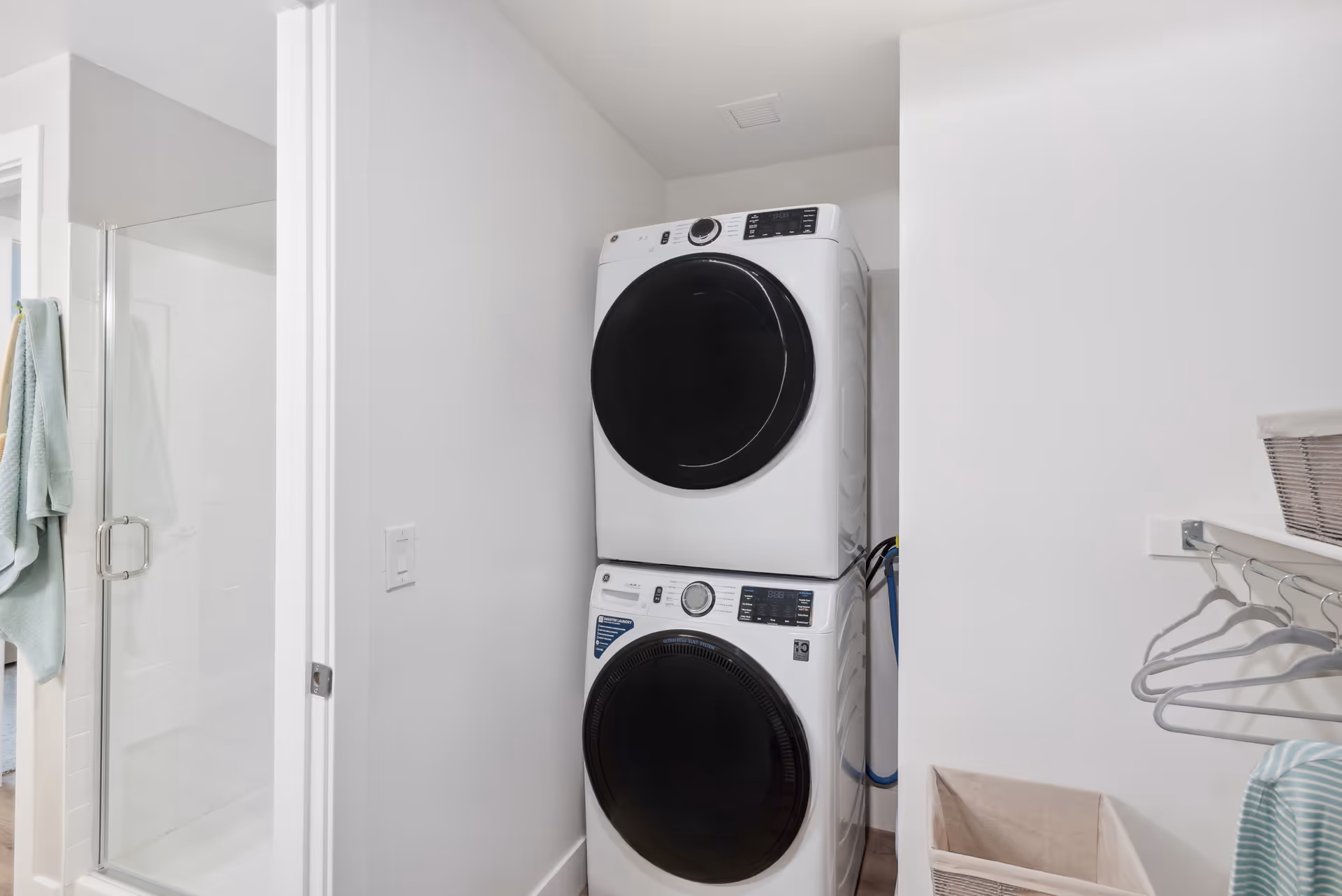 Stacked front-loading washer and dryer in a small laundry nook next to a bathroom shower with towels and hanging shelves.