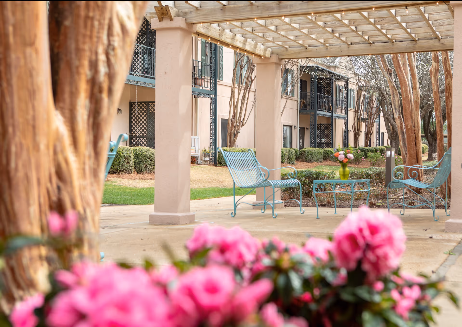 Outdoor seating area at The Orchard with light blue metal chairs and a small table holding a green vase with flowers, under a wooden pergola. Pink flowers and tree trunks are visible in the foreground, with a beige building and trimmed bushes in the background.
