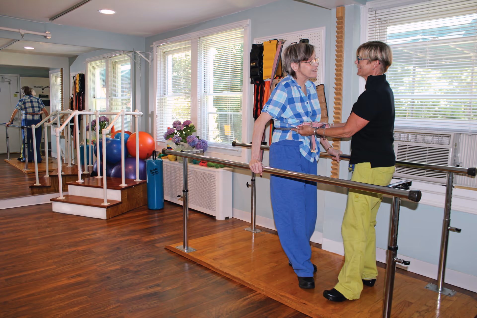 A senior woman wearing a blue plaid shirt and blue pants is assisted by a caregiver dressed in a black top and yellow pants while walking between parallel bars in a physical therapy or rehabilitation room. The room has wooden floors, large windows with blinds, exercise balls, and a mirrored wall reflecting the scene.