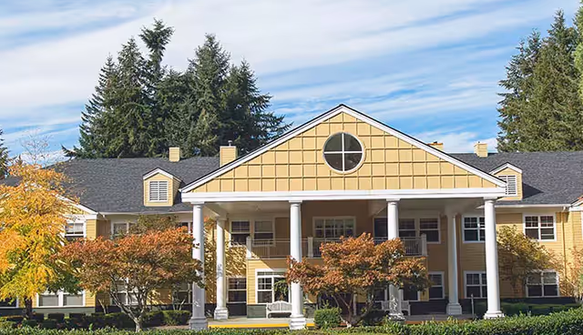 Yellow two-story assisted living building with a covered porch supported by white columns and landscaped trees in front.