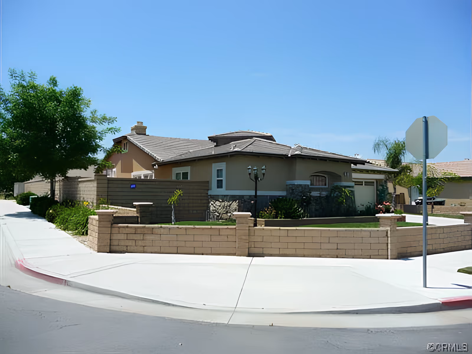 Exterior view of a single-story residential building with a beige facade, stone accents, and a tiled roof. The building is surrounded by a low brick wall and has a small lawn area with some plants and a tree. A sidewalk and a street corner with a stop sign are visible in the foreground under a clear blue sky.