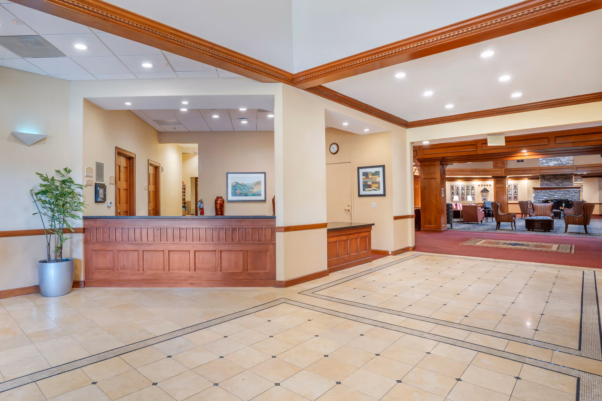 Interior view of a senior living facility lobby with a wooden reception desk, beige tiled floor, cream-colored walls, and recessed ceiling lights. There is a potted plant on the left, framed artwork on the walls, and an open doorway leading to a lounge area with chairs and a stone fireplace.