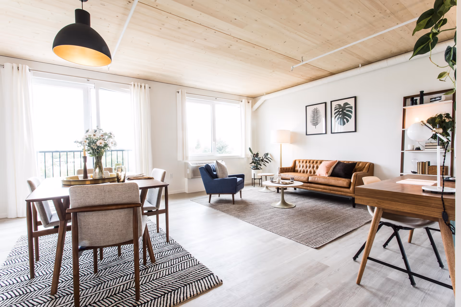 A modern living room featuring a brown leather sofa, a dining table with chairs, and large windows allowing natural light. The decor includes plants and framed artwork on the walls.