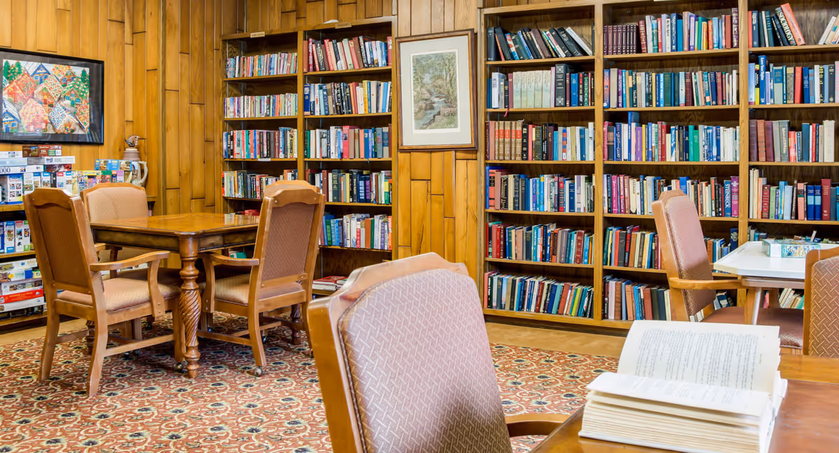A cozy library reading area featuring wooden bookshelves filled with books, a wooden table surrounded by chairs, and a patterned rug.