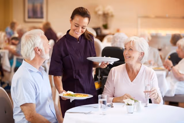 A female caregiver serving food to an elderly man and woman seated at a dining table in a communal dining room. The elderly woman is holding a glass of water and smiling at the caregiver. Other elderly people are visible in the background, also seated at tables.