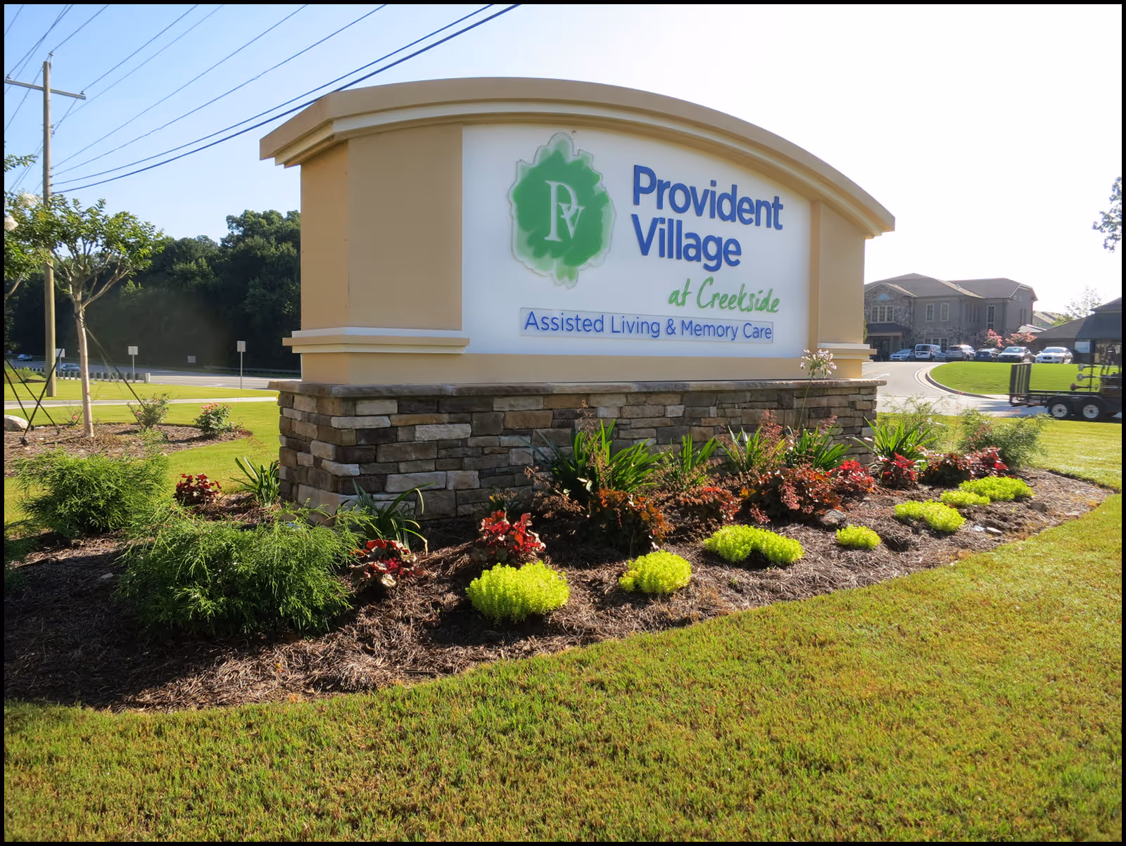Outdoor view of a large sign for Provident Village at Creekside Assisted Living & Memory Care, surrounded by landscaped greenery and flowers, with a building and driveway visible in the background under a clear sky.