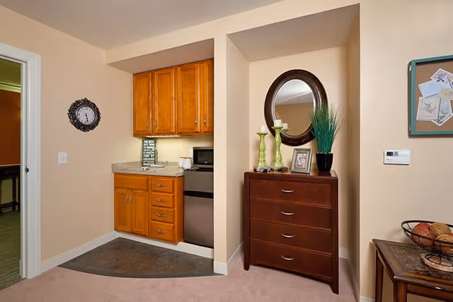 Small kitchenette area with wooden cabinets, a mini fridge, microwave, and countertop. Adjacent to the kitchenette is a dark wooden chest of drawers with decorative items including two green candlesticks, a framed photo, a potted plant, and a round mirror above it. A wall clock is mounted on the left wall, and a bulletin board with pinned papers is on the right wall.