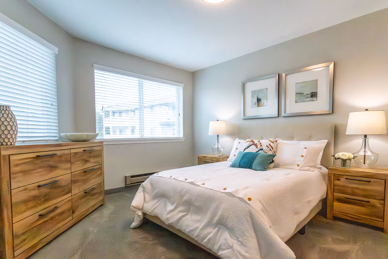 A bright and cozy bedroom with a neatly made bed featuring white bedding and decorative pillows. The room has two wooden nightstands with lamps on each side of the bed, a wooden dresser with a decorative bowl and vase, and two framed pictures hanging above the bed. Large windows with white blinds allow natural light to fill the room.