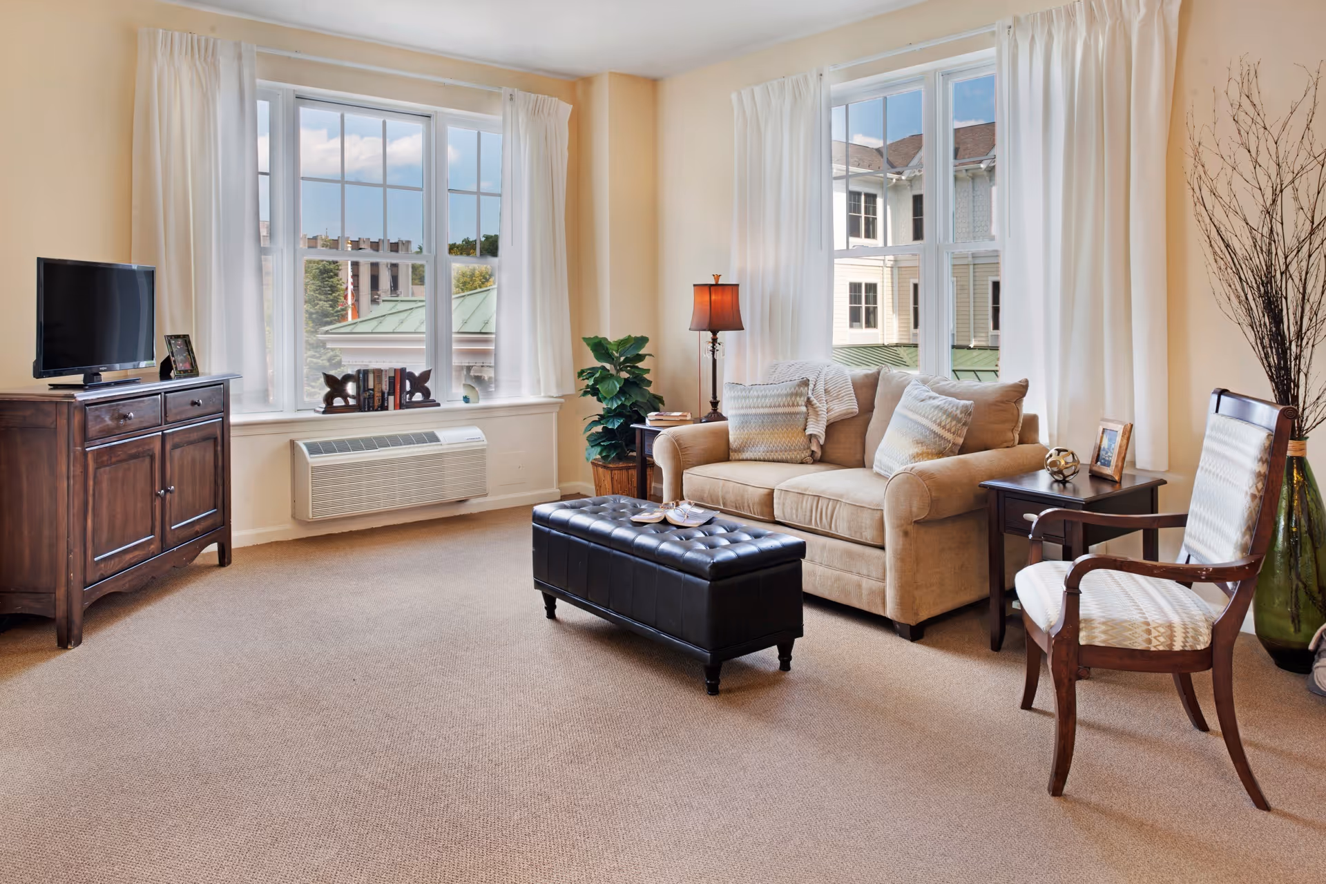 A cozy living room with beige walls and carpet, featuring a beige sofa with patterned pillows, a dark wooden side table with a framed photo and decorative item, a floor lamp, a wooden armchair with patterned upholstery, a black tufted leather ottoman, a wooden cabinet with a flat-screen TV and a small framed picture, and large windows with white curtains letting in natural light.