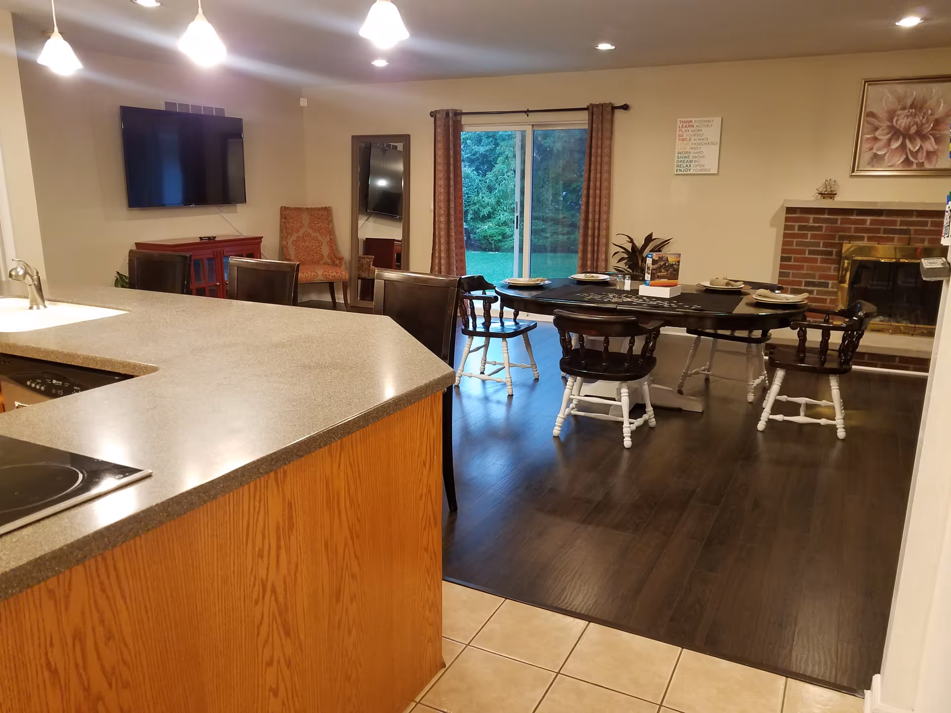 Interior view of a senior living facility dining and living area at Ann Cook Center. The image shows a kitchen counter with a sink in the foreground, a dining table set with plates and chairs on a dark wood floor, a brick fireplace with a floral painting above it, a sliding glass door with curtains leading outside, a wall-mounted TV, an upholstered chair, and a full-length mirror.
