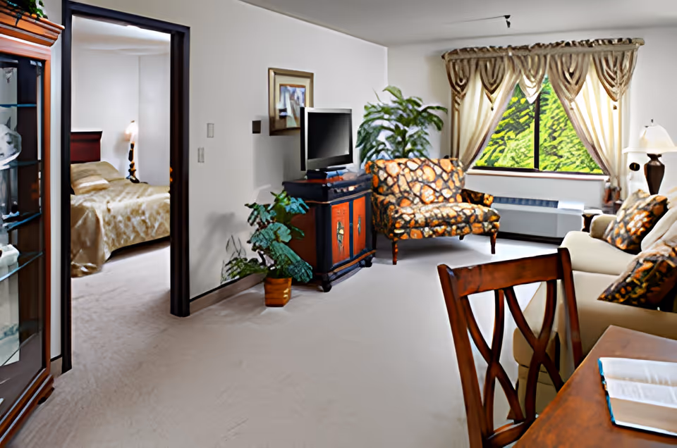 A cozy living room area with a patterned loveseat and matching pillows on a beige sofa, a wooden TV stand with a flat-screen TV, potted plants, and a window with decorative curtains letting in natural light. To the left, an open doorway reveals a bedroom with a bed covered in a gold bedspread and a bedside lamp.