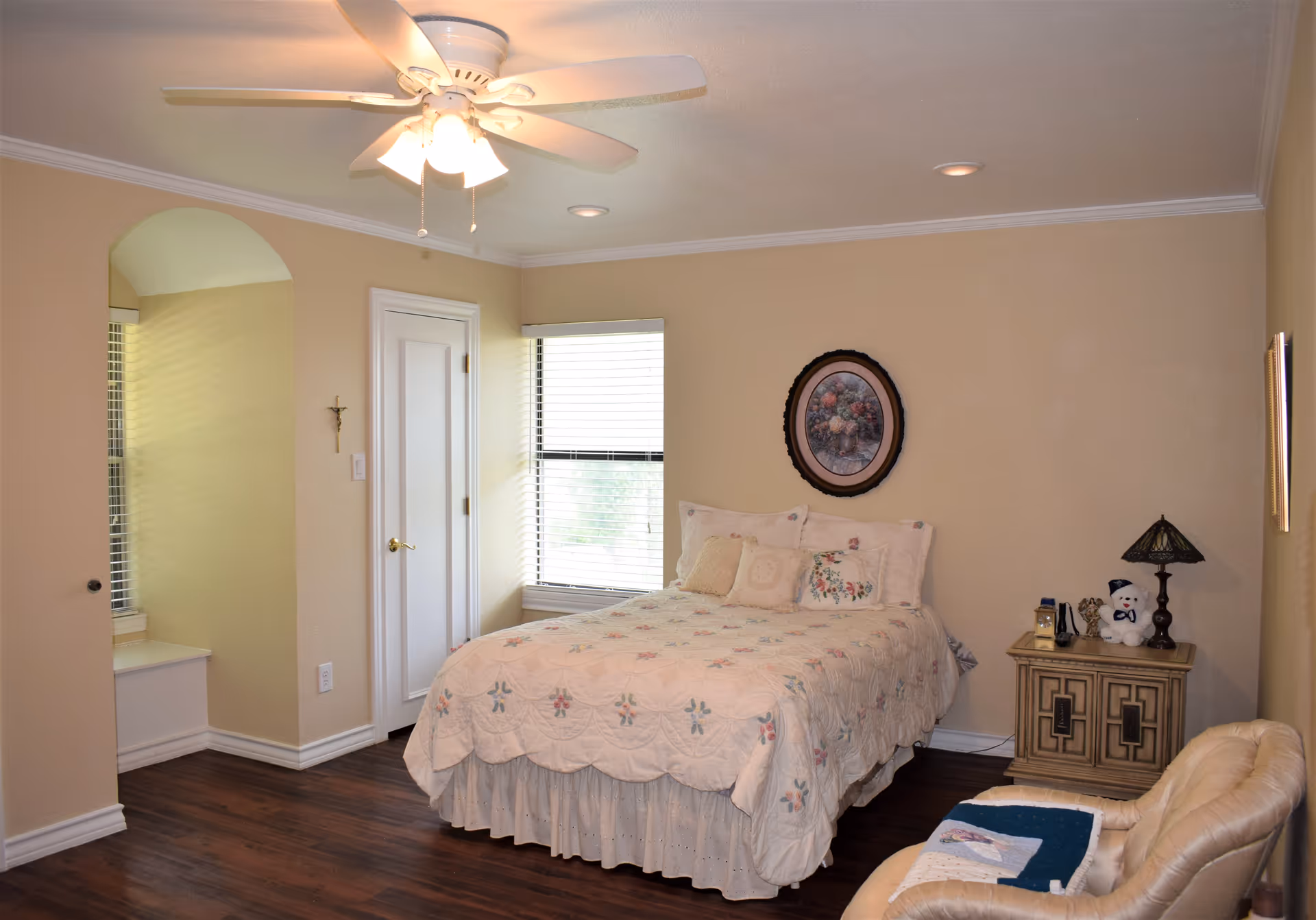 A cozy bedroom with a bed covered in a floral quilt, a nightstand with a lamp and decorative items, a beige armchair with a quilt draped over it, a window with blinds, a ceiling fan with lights, and light beige walls with a framed floral picture above the bed.