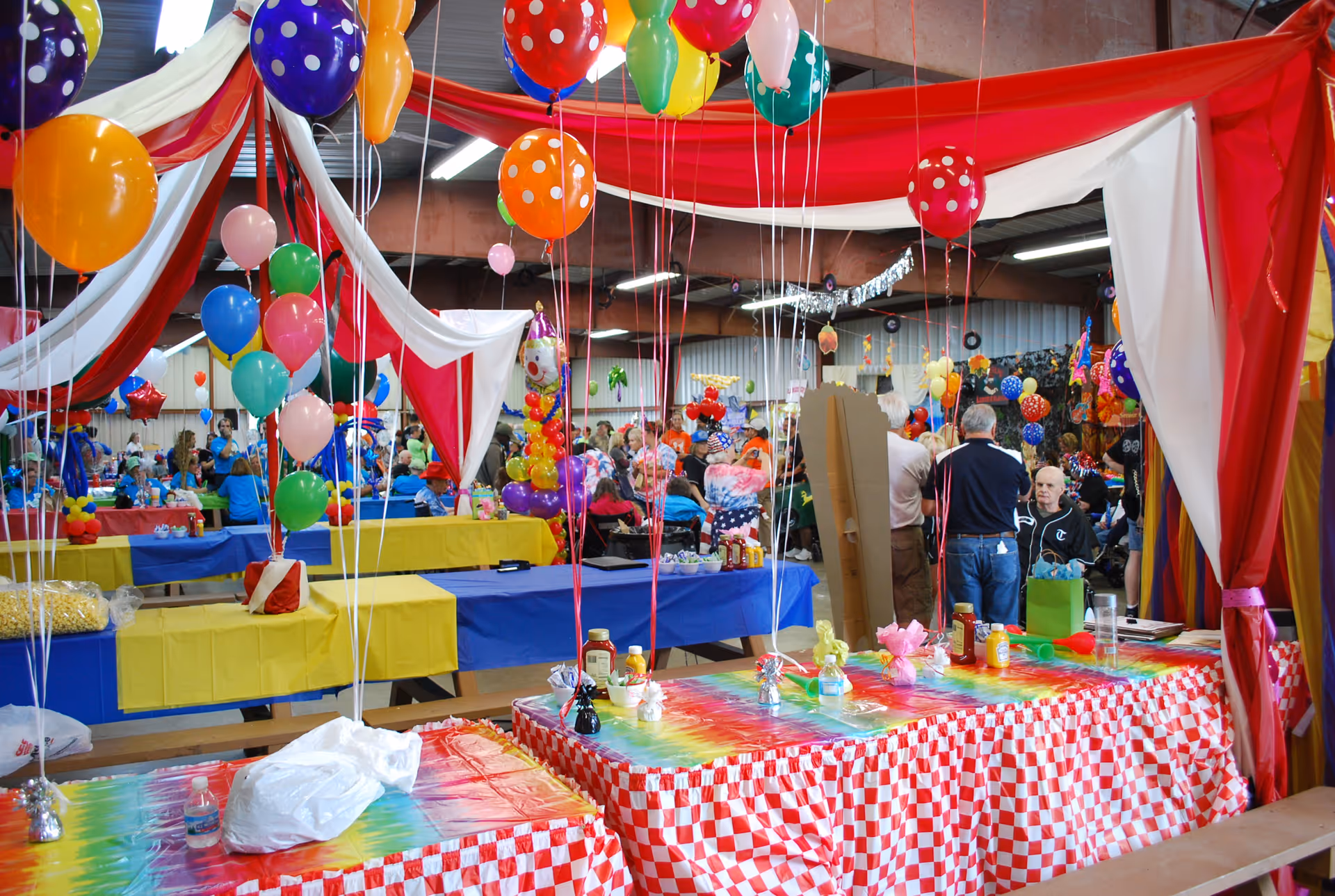 A large indoor party or event space decorated with colorful balloons, red and white draped fabric, and tables covered with bright tablecloths in red checkered and tie-dye patterns. People are gathered in the background, sitting and standing, enjoying the festive atmosphere.