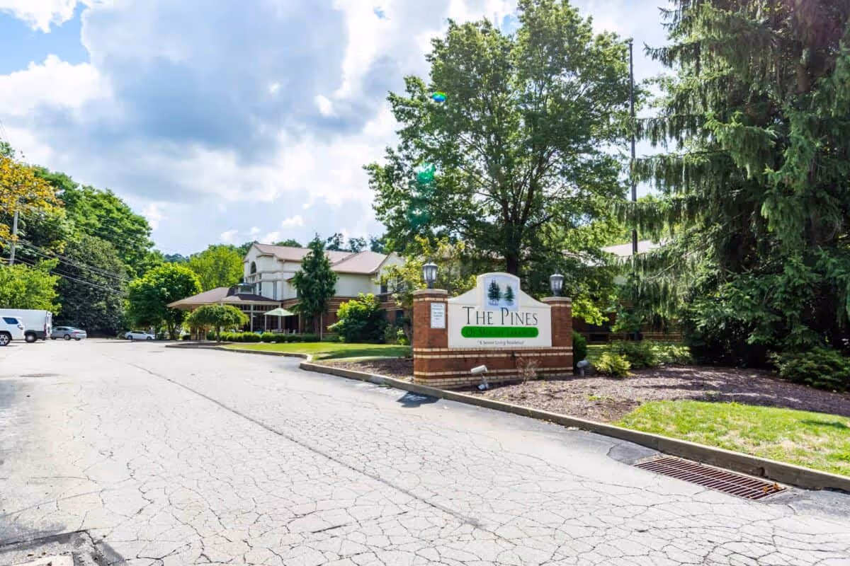 Driveway and entrance to The Pines senior living facility with a brick sign, building façade, and surrounding trees.