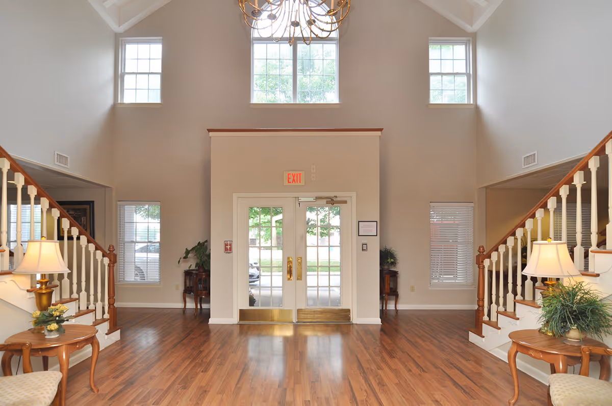 Interior view of a spacious senior living facility entrance with double glass doors, wooden flooring, two symmetrical staircases with white railings on either side, and large windows letting in natural light. There are small tables with lamps and plants near the staircases.