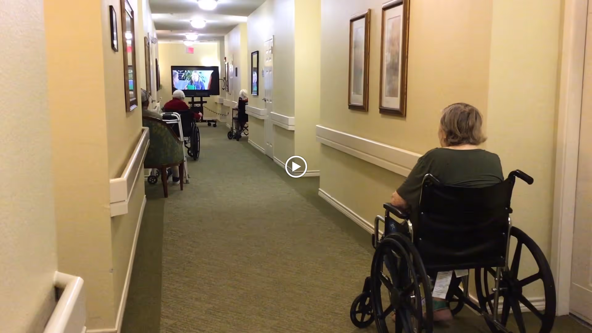 A hallway in a senior living facility with beige walls and carpeted floor. Several elderly individuals are seated in wheelchairs along the hallway, facing a television screen mounted at the far end. The walls have handrails and framed pictures.