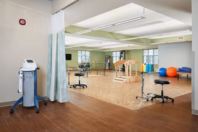 Spacious senior living rehabilitation room with therapy equipment, parallel bars, exercise balls, stools, and practice stairs.