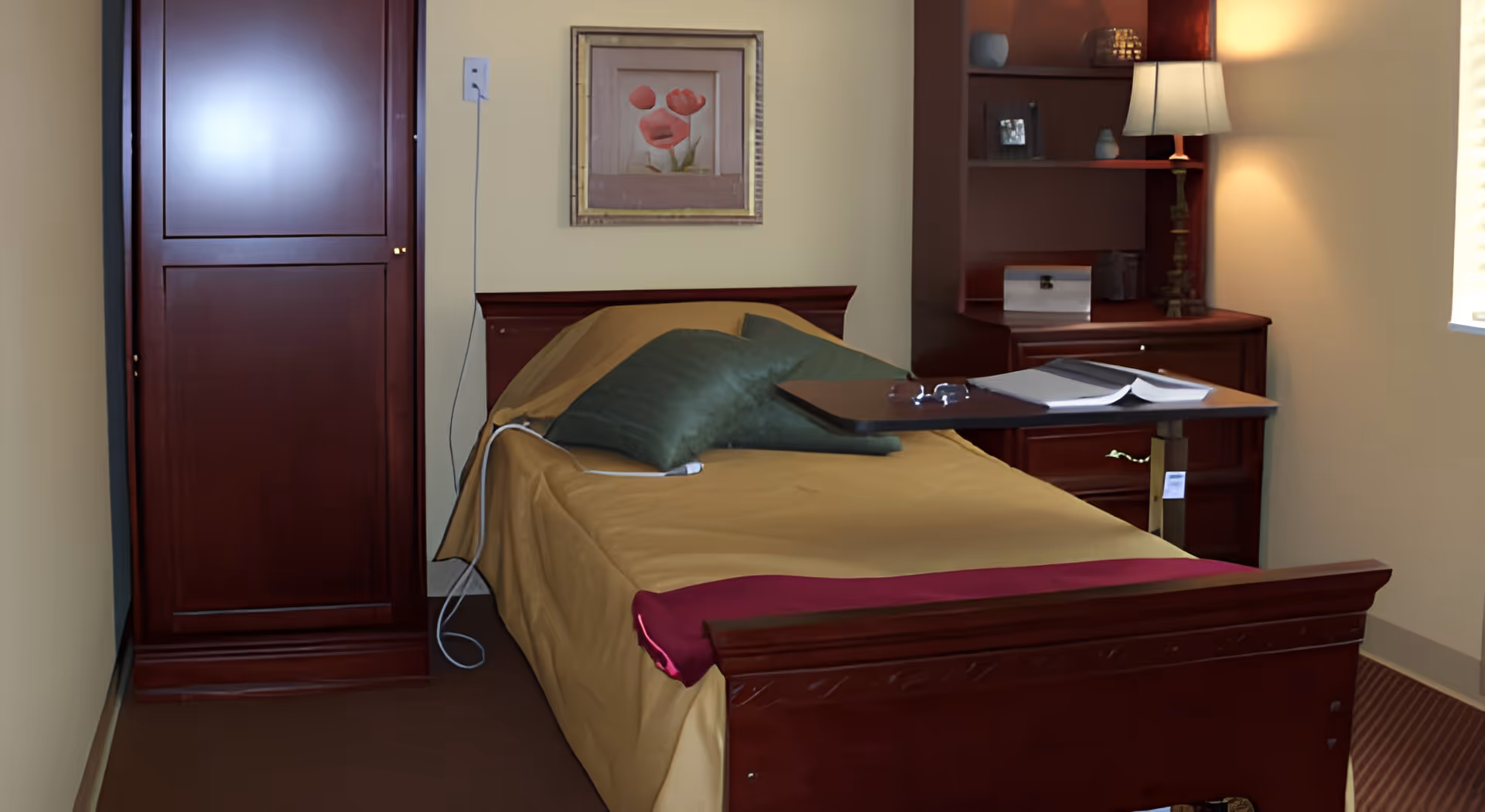 A furnished bedroom with a single bed, wooden wardrobe and nightstand, lamp, overbed table, and framed art on the wall.