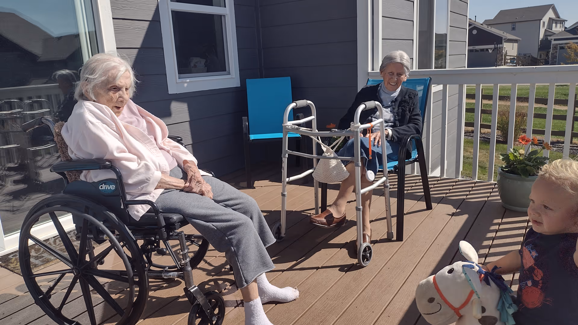 Two elderly women — one in a wheelchair and one with a walker — sit on a sunny deck while a small child plays with a toy horse nearby.
