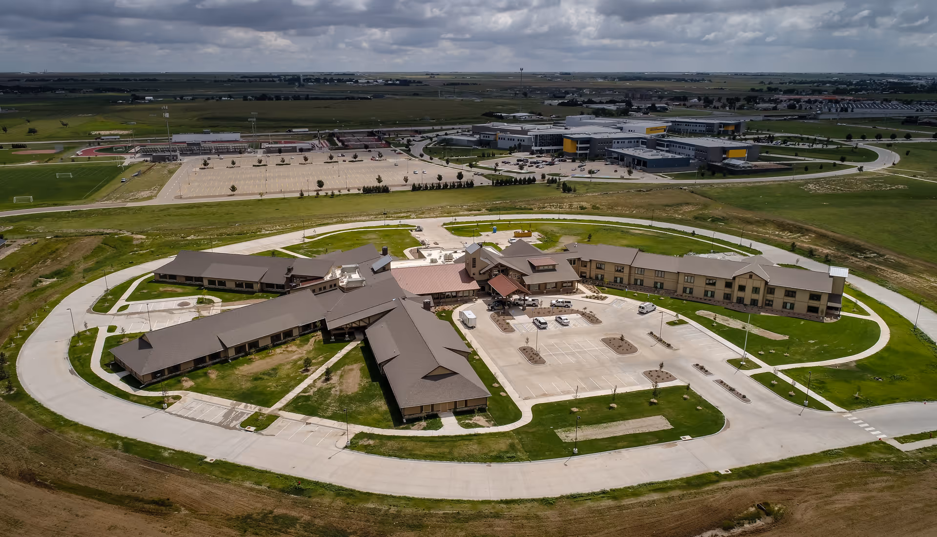 Aerial view of Ranch House Senior Living facility showing a large, single-story building with multiple wings, surrounded by a circular driveway and parking areas. The building is set in a green landscape with open fields and other buildings visible in the distance under a cloudy sky.
