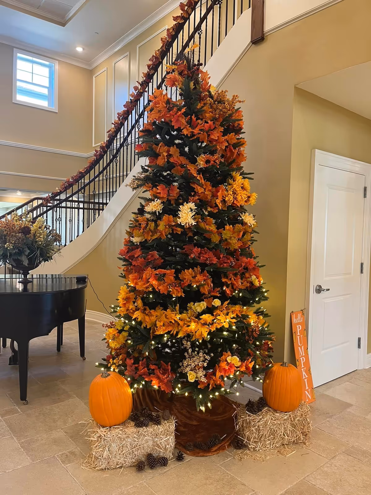 A decorated artificial tree with autumn-themed foliage and flowers in shades of orange, yellow, and red, adorned with small white lights. The tree is placed indoors near a staircase with a black railing wrapped in matching autumn garland. Two pumpkins sit on hay bales at the base of the tree, along with pinecones scattered around. A sign next to the tree reads 'hello PUMPKIN'. A black grand piano is visible to the left, and a white door is on the right side of the image.