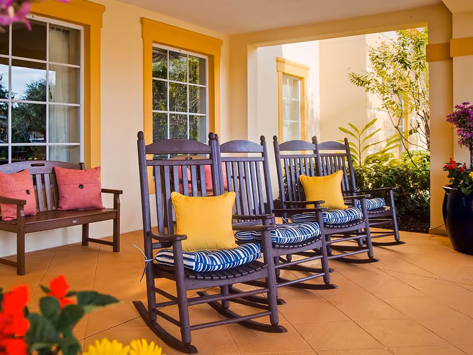 A covered porch with wooden rocking chairs and cushions, a bench with pillows, and potted plants.