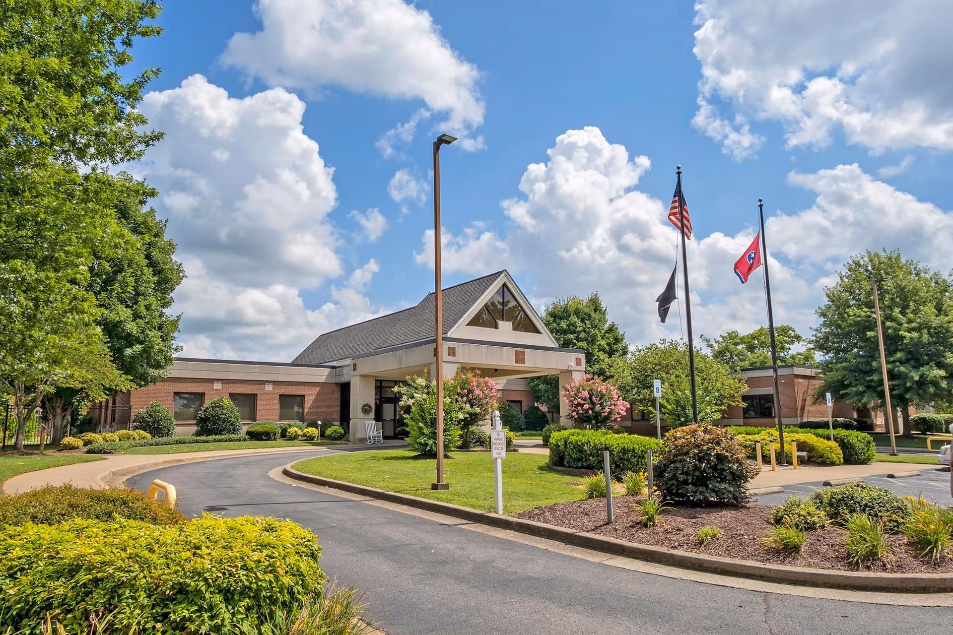 Exterior view of the TN State Veterans Home building with a curved driveway, landscaped bushes, trees, and three flagpoles flying the American flag, a black flag, and the Tennessee state flag under a partly cloudy blue sky.