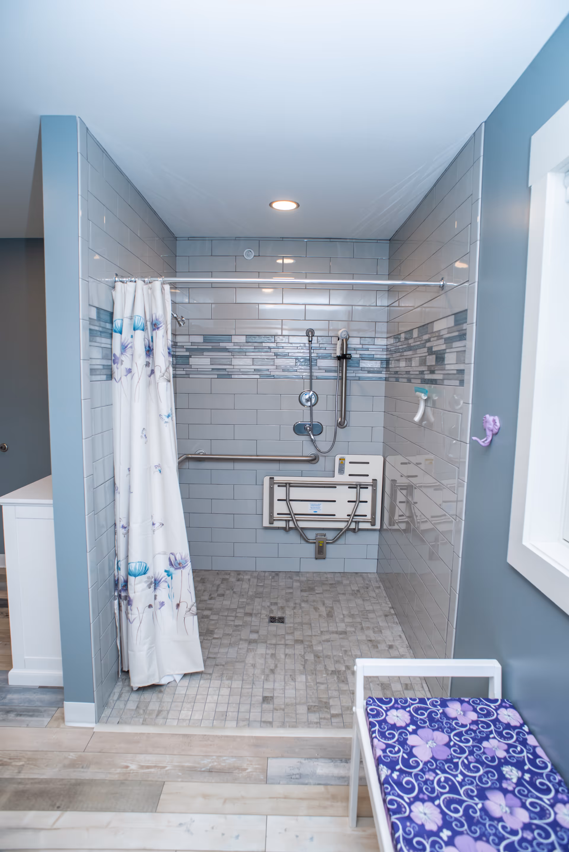 A modern accessible shower area with gray tiled walls and floor, featuring a fold-down shower seat, grab bars, a handheld showerhead, and a floral shower curtain. A small bench with a purple floral cushion is positioned outside the shower area.