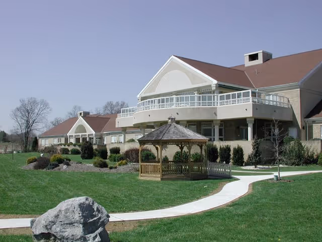 Exterior view of Diakon Senior Living Ravenwood Campus showing a large building with a brown roof and beige walls. In front of the building is a well-maintained lawn with a wooden gazebo and a curved concrete walkway. Trees and shrubs are planted around the building and lawn area under a clear blue sky.