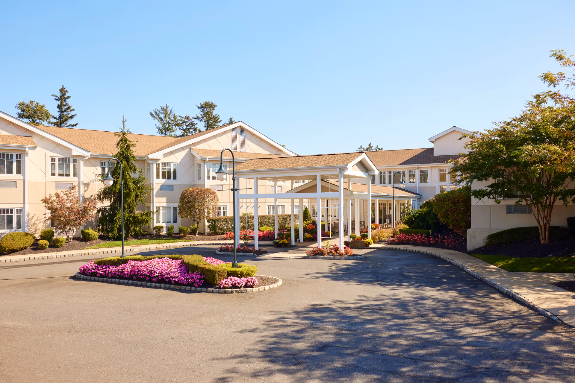 Exterior view of Brandywine Governor's Crossing by Monarch senior living facility showing a large building with beige walls and a brown roof. The entrance has a covered driveway with white pillars, surrounded by well-maintained landscaping including green shrubs, trees, and vibrant pink flowers under a clear blue sky.