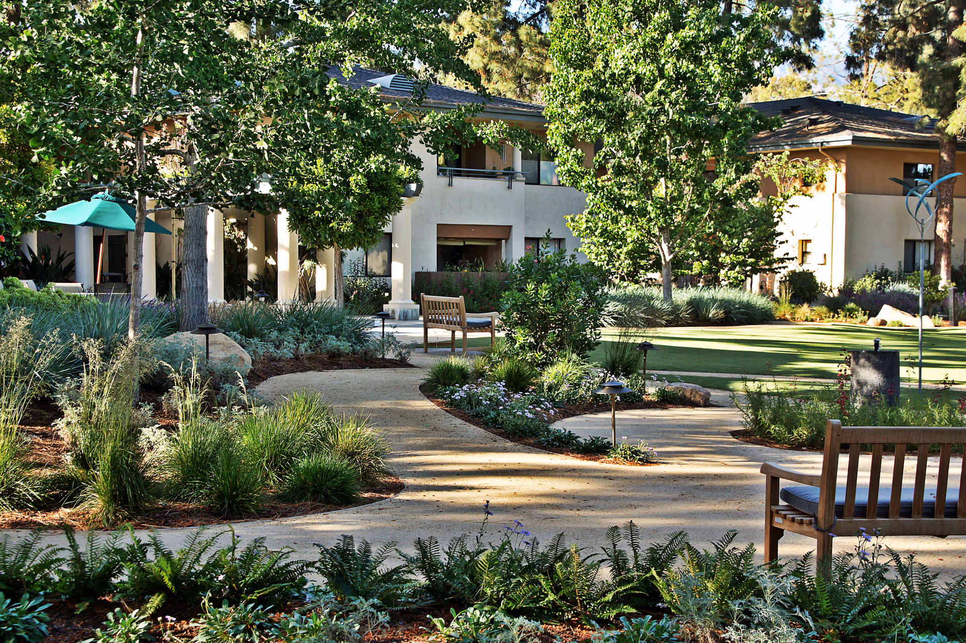A landscaped outdoor garden area at Vista del Monte Retirement Community featuring a winding paved pathway, green grass, various trees and shrubs, wooden benches, and a building in the background with balconies and shaded patio areas.