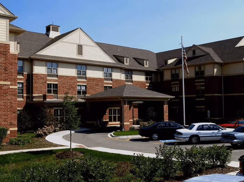 Exterior view of a multi-story brick and beige senior living facility building with a covered entrance, several parked cars, a flagpole with an American flag, and landscaped greenery in front.