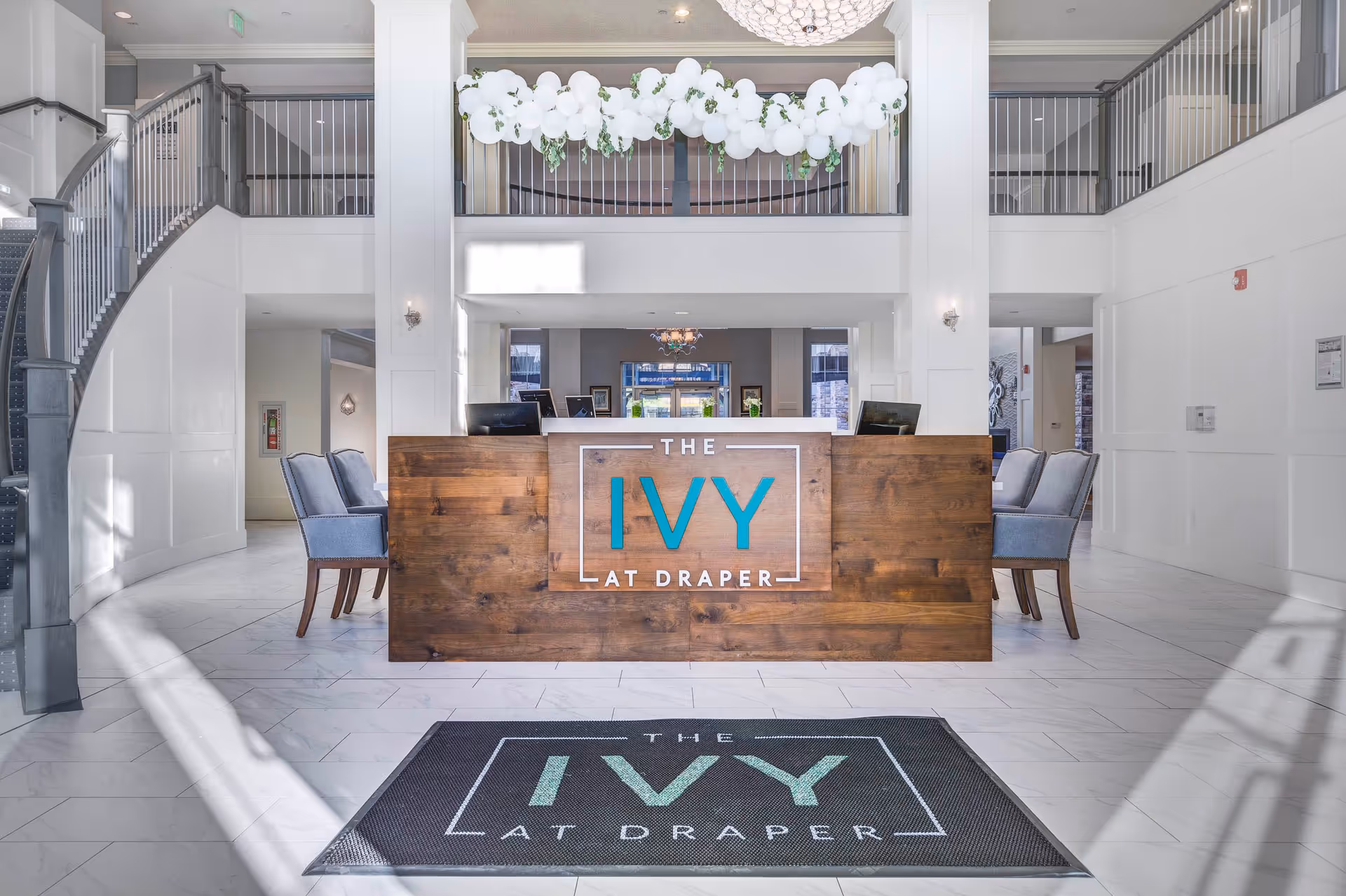 Bright and spacious reception area of The Ivy at Draper featuring a wooden front desk with the facility's name displayed, a decorative white balloon arrangement above, two blue upholstered chairs on each side, and a staircase with metal railings on the left side.