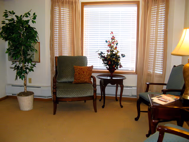 A cozy living room area with two upholstered armchairs, a small round wooden table with a floral arrangement, a potted plant in the corner, and a window with beige curtains and blinds letting in natural light. A table lamp and some magazines are visible on a side table next to one of the chairs.