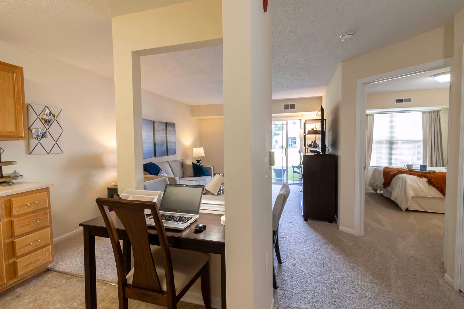 Interior view of an apartment at Independence Village of White Lake showing a small workspace with a laptop and chair, a living room with a couch and lamp, and a bedroom with a bed and window. The space is well-lit with natural light coming through sliding glass doors and windows.