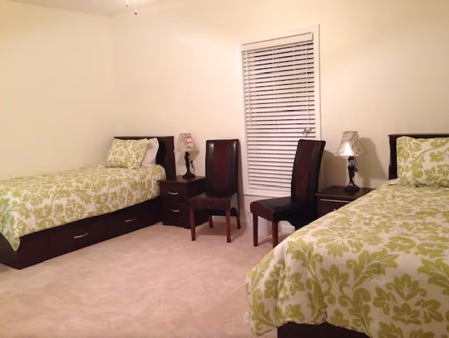 A simple bedroom with two twin beds dressed in green patterned bedding, nightstands, chairs, and a window with closed blinds.