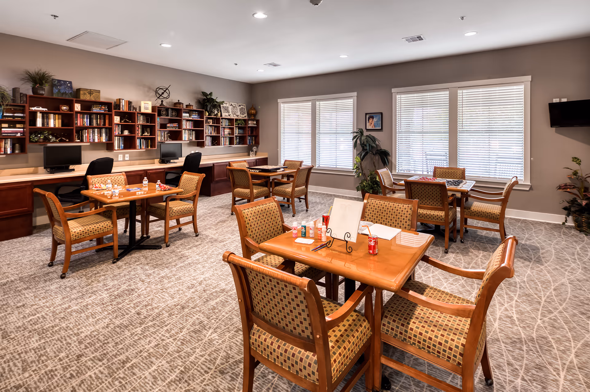 A well-lit common room with several wooden tables and cushioned chairs arranged for group activities or dining. The room features a carpeted floor, large windows with blinds, and a wall with built-in bookshelves filled with books and decorative items. There are two computer workstations along the back wall and some plants placed around the room.