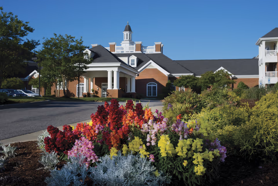 A senior living facility building with a brick exterior and white columns at the entrance, surrounded by well-maintained landscaping including colorful flowers and green shrubs under a clear blue sky.