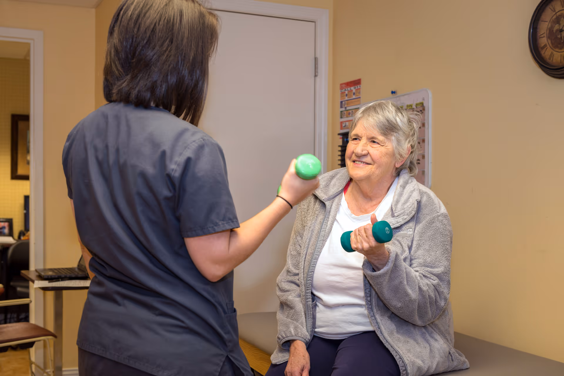 An elderly woman sitting on a bench in a room, smiling and holding a green dumbbell in her right hand, while a caregiver or physical therapist in gray scrubs stands in front of her holding a similar green dumbbell.