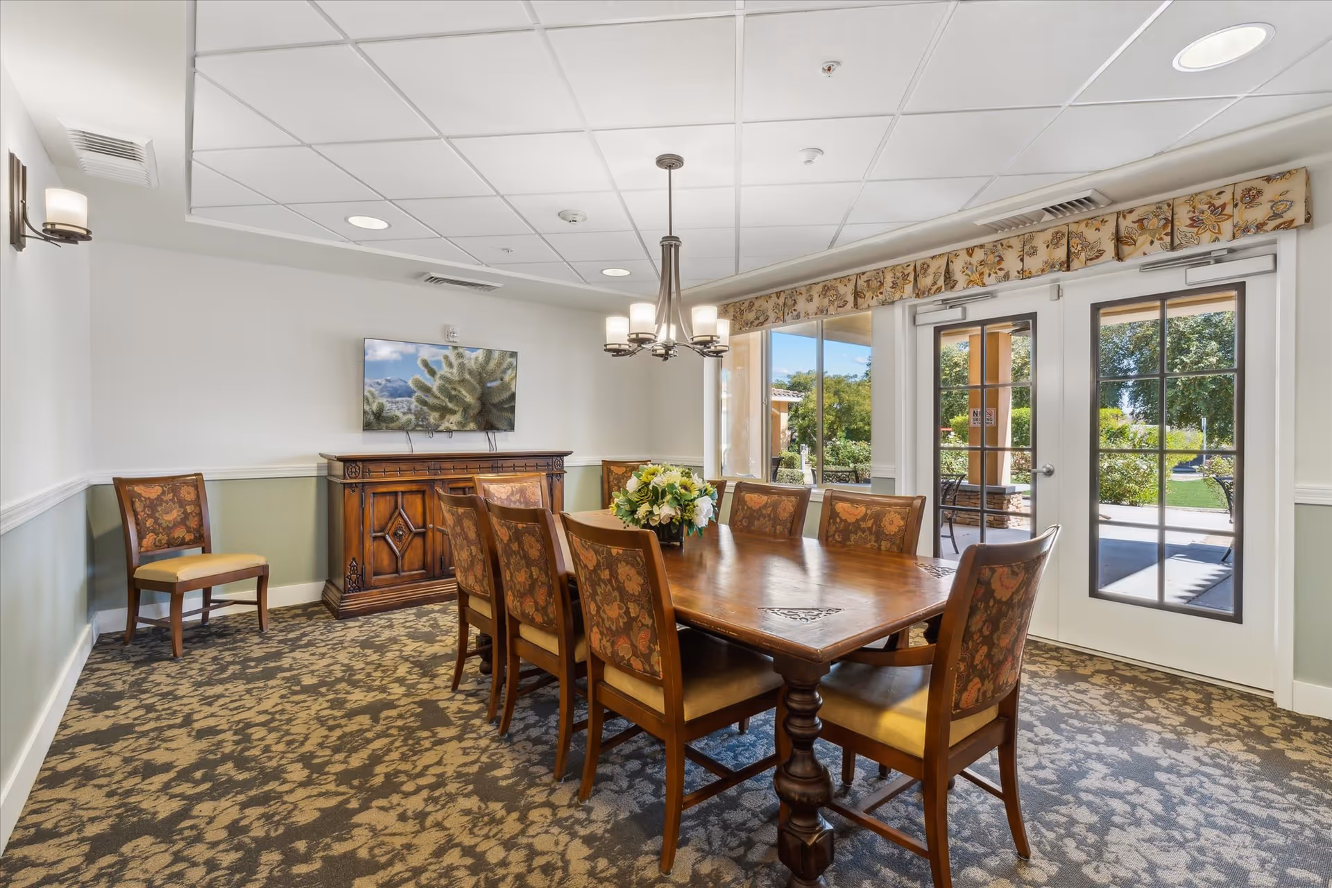 A well-lit dining room with a wooden dining table surrounded by eight upholstered chairs with floral patterns. A floral centerpiece is on the table. The room has patterned carpet flooring, light green and white walls, and a ceiling with recessed lighting and a chandelier. A wooden cabinet with a flat-screen TV mounted above it is against one wall. French doors with glass panes open to an outdoor garden area with greenery and seating.