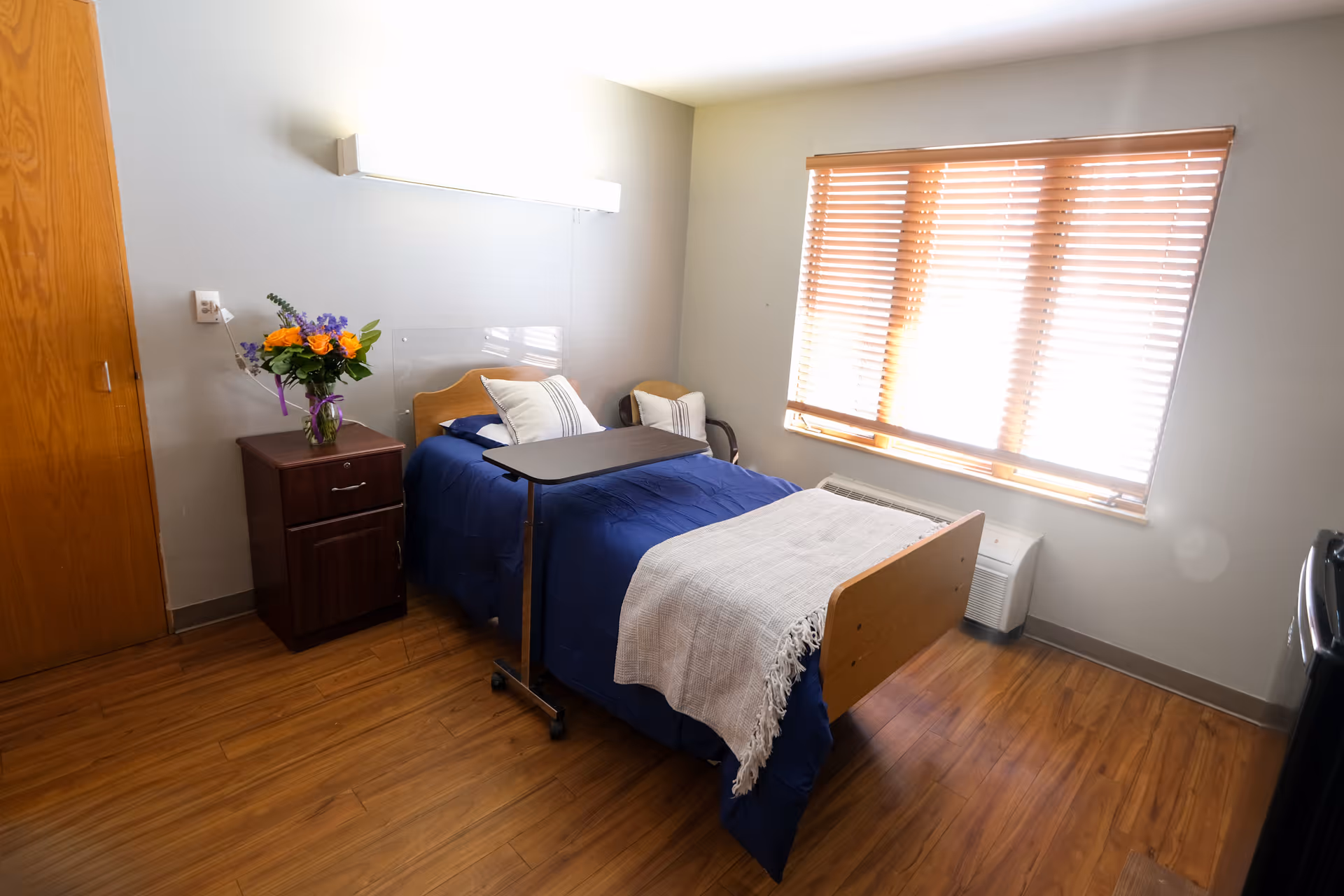 A small, tidy bedroom with a single bed covered in a blue blanket and a white throw. There is a wooden nightstand with a vase of colorful flowers next to the bed. A window with wooden blinds allows natural light into the room. The floor is wooden, and there is a small overbed table positioned over the bed.
