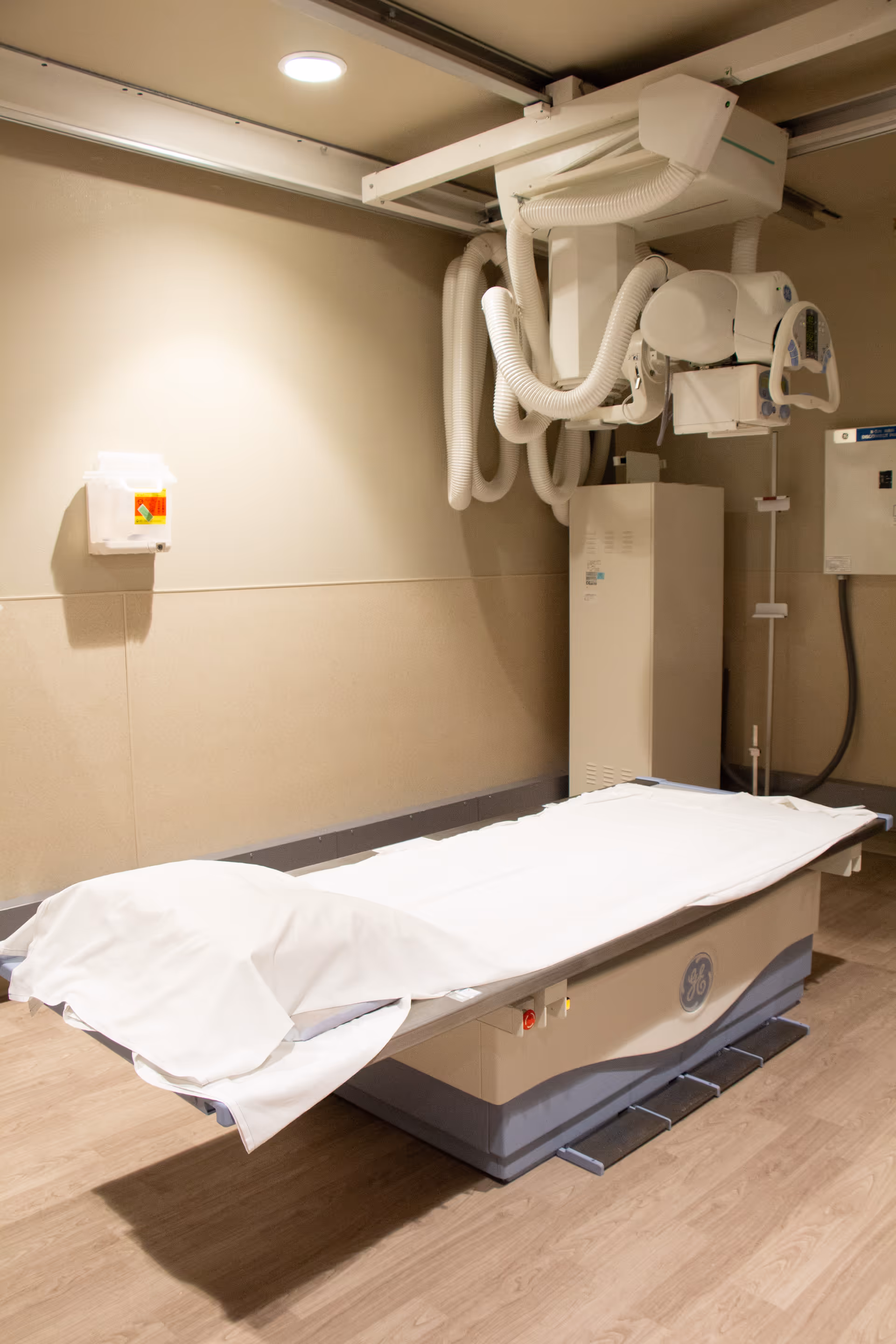 A medical imaging room with an X-ray machine mounted on the ceiling and a patient examination table covered with a white sheet. The room has beige walls and wood-patterned flooring.