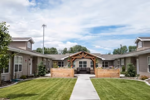 Exterior view of The Gables Assisted Living of Idaho Falls showing a single-story building with a central covered patio area, surrounded by well-maintained green lawns and trees under a partly cloudy sky.