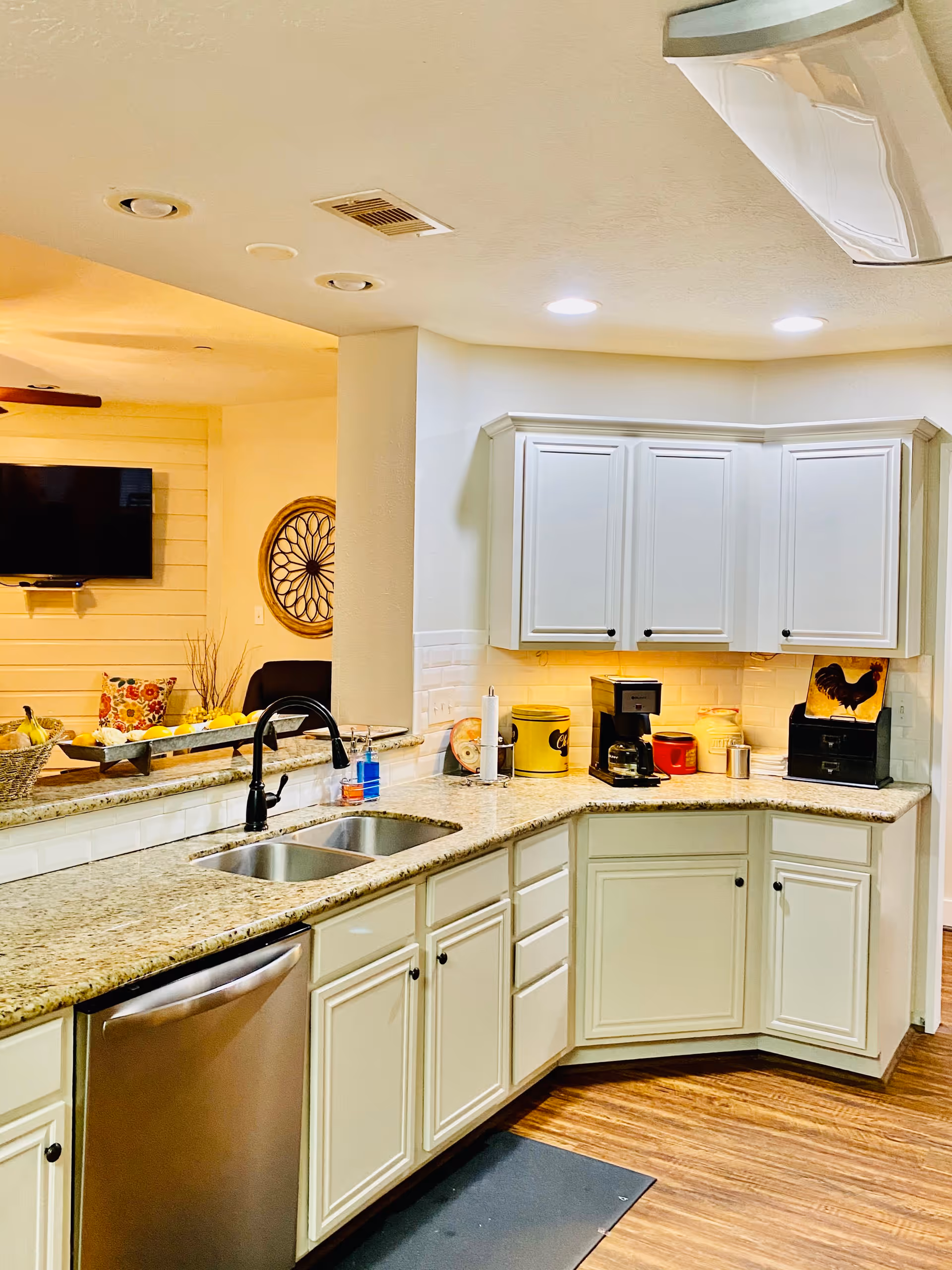 A bright kitchen with white cabinets, granite countertops, and a double sink. There is a stainless steel dishwasher below the counter. On the countertop, there is a coffee maker, a paper towel holder, and various kitchen containers. The kitchen opens to a living area with a wall-mounted TV and decorative items.
