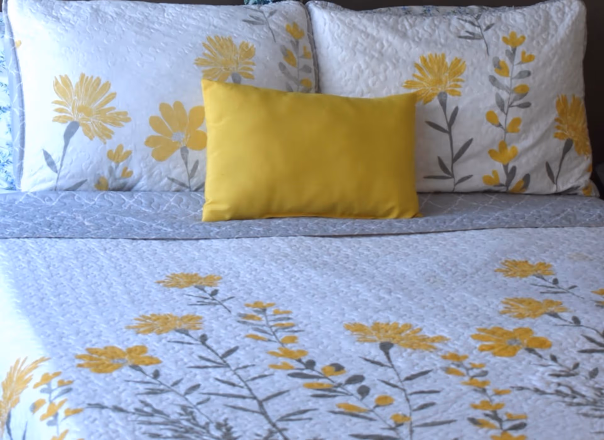 A neatly made bed with white and yellow floral-patterned bedding and pillows, including a solid yellow rectangular accent pillow in the center.