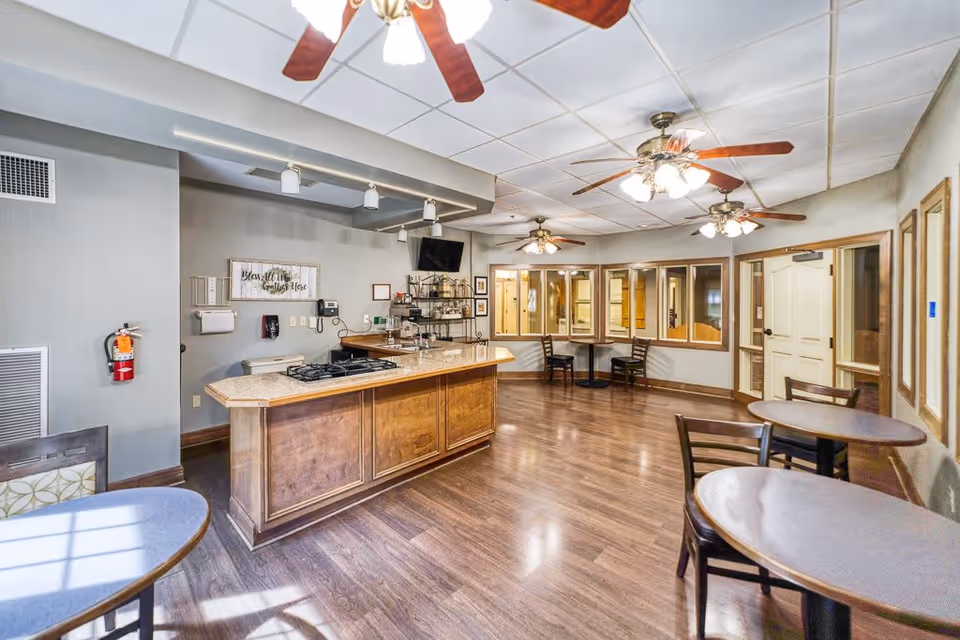 Interior view of a common area in a senior living facility featuring a kitchen island with a stove, several round tables with chairs, ceiling fans with lights, and a wall-mounted TV. The room has wooden flooring, light gray walls, and multiple windows looking into adjacent rooms.