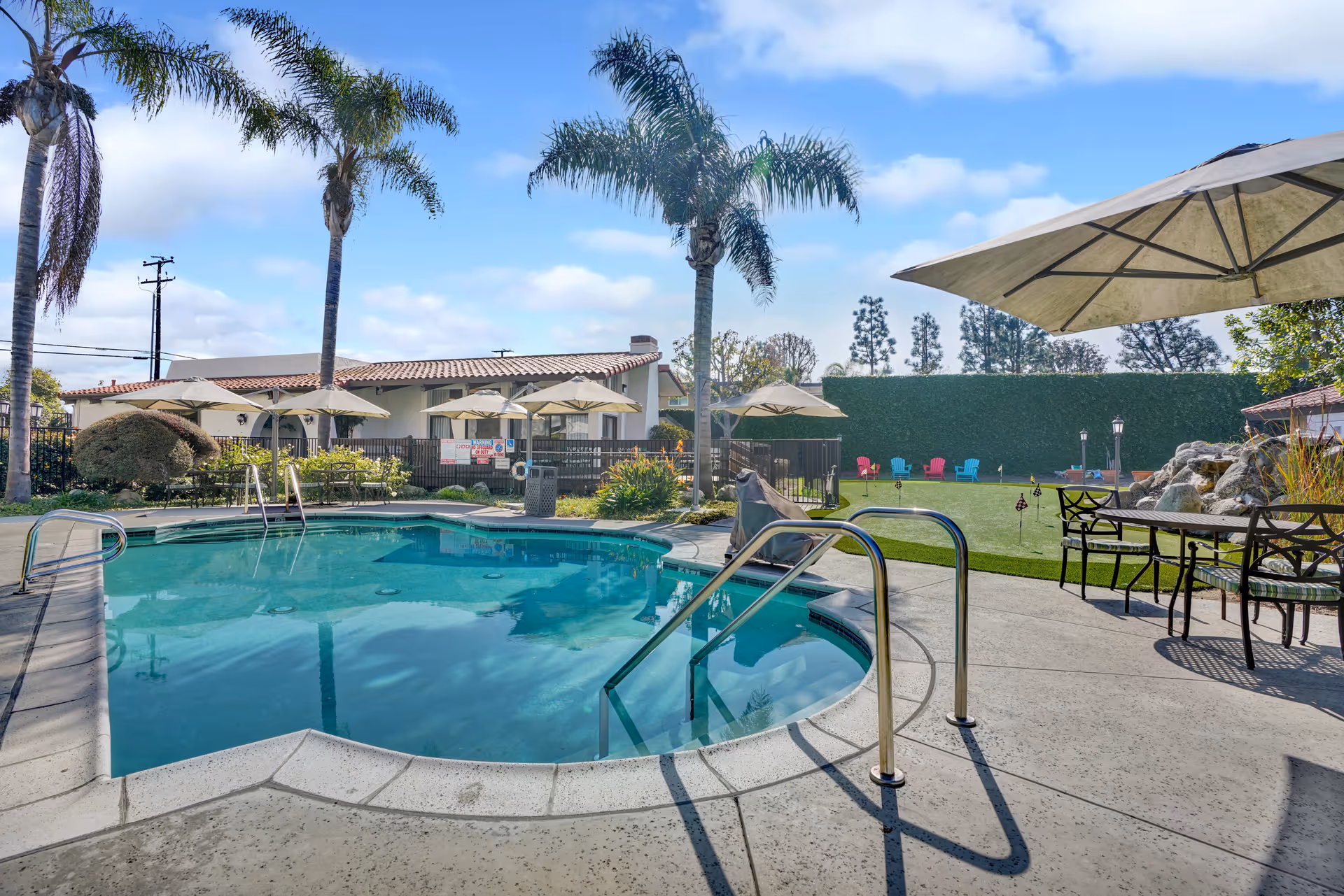 Outdoor swimming pool area with palm trees, patio tables with umbrellas, and colorful chairs on a grassy lawn under a blue sky with some clouds.